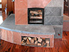 Firewood is loaded and ready to burn in this 6 KW Soapstone/Sandstone masonry heater.  Curved benches and a wide mantle grace this asymmetrical masterpiece.  Though not obvious here, this is a two-story heater culminating in a soapstone bench upstairs.  The original exterior design of this masonry heater is by architect Martin Johannessen at Harmoni Designs.